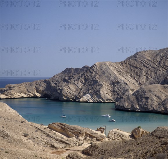 OMAN, Capital Area, Bander Jissah, "Gulf of Oman. View from coastline over bay with yachts and a schooner on calm blue sea, cliffs behind."