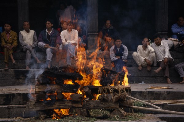 NEPAL, Kathmandu, Hindu cremation in Pashupatinath Temple.  Burning wood funeral pyre with onlookers on steps behind.