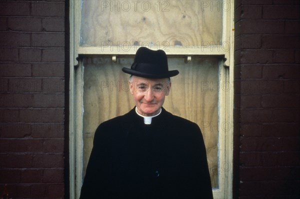 IRELAND,  , Dublin, Priest wearing hat in front of boarded up house window.
