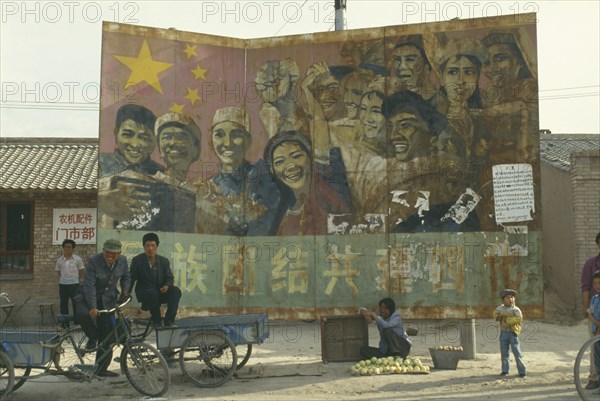CHINA, Ningxia, Guyuan , Old Communist poster on hoarding at road junction with woman selling fruit below. Child and two men on stationary bicycle driven carts.