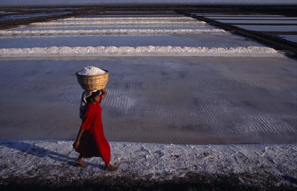 10127776 INDIA Gujarat Diu Woman wearing red carries salt in basket on her head across white salt pan