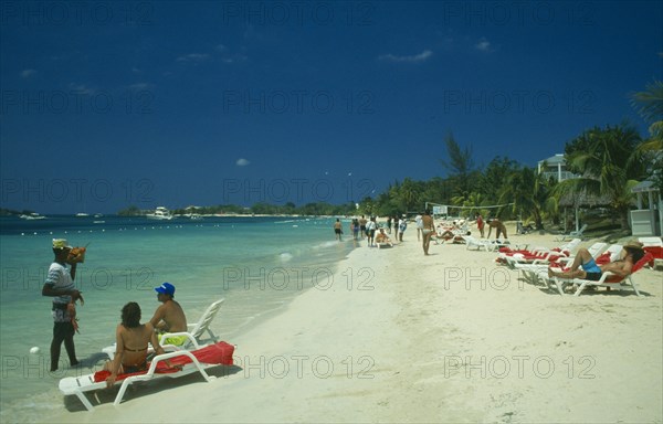 WEST INDIES, Jamaica, Negril, Man selling lobster to tourists on beach sitting on sun loungers by the water with other tourist sunbathing