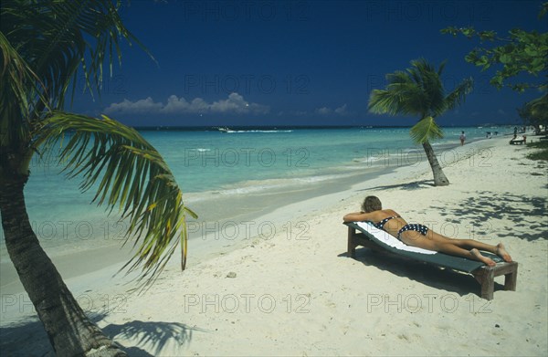 WEST INDIES, Jamaica, Negril, Woman sunbathing on lounger on beach near water and coconut palm trees