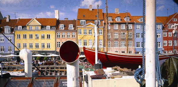 DENMARK, Copenhagen, Nyhavn Canal. View over boats to pavement cafes and waterside buildings.