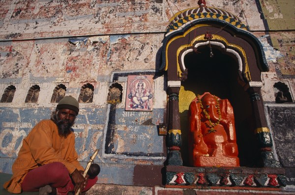 INDIA, Maharashtra, Nasik, Sadhu sitting beside shrine dedicated to Ganesh with orange painted figure of elephant headed god draped with garland of marigold flowers.