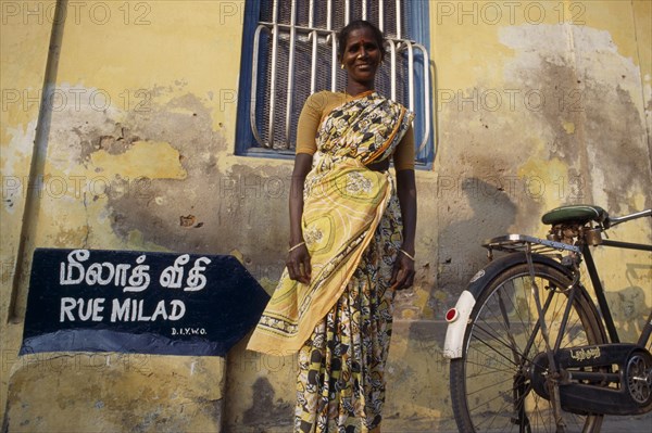 INDIA, Tamil Nadu, Pondicherry, Woman in a yellow sari standing in front of a yellow wall beside a bicycle.