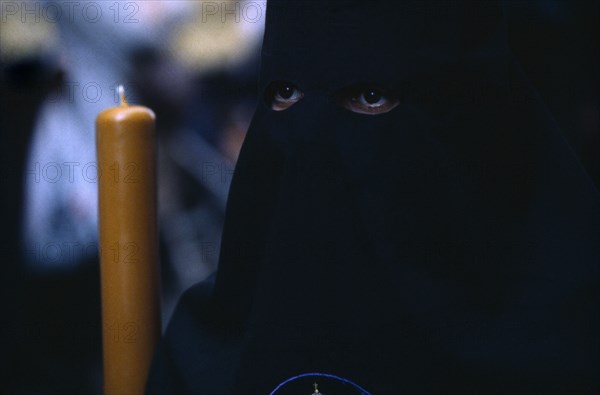 SPAIN, Andalucia, Seville, Semana Santa Easter procession. Penitent in black robe with eyes visable through hood holding a candle