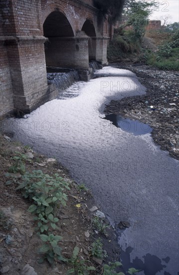 INDIA, Uttar Pradesh, Agra , Polluted river.  Scum and foam covered water flowing through arches in bridge and leaving wake of rubbish and other debris.