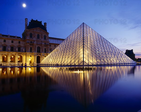FRANCE, Ile de France, Paris, Louvre.  Glass pyramid and surrounding buildings floodlit at night and reflected in water in the foreground.