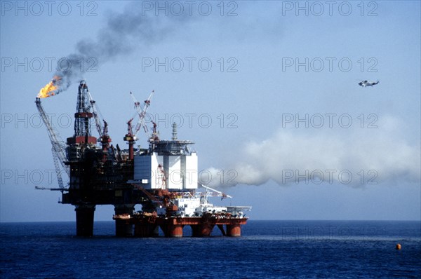 INDUSTRY, Factory, Oil Rig, Worker in a hard hat standing on the deck watching flames from burning off gas over the North sea