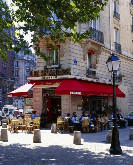 FRANCE, Ile de France, Paris, Cafe on street corner with red awning and outside tables street lamp and tree.