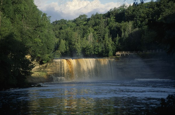 USA, Michigan , Chippewa, Tahquamenon Falls waterfall surrounded by lush greenery and man standing on riverbank nearby