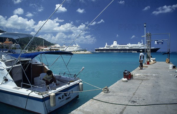 WEST INDIES, Jamaica, Ocho Rios, Harbour view with man standing on stone jetty and cruise ships behind.  Moored boat in the foreground with person sitting in shade on deck.