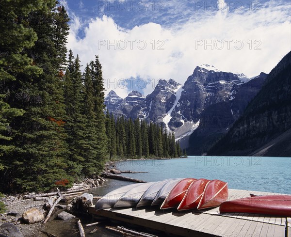 CANADA, Alberta, Moraine Lake, Canoes on a jetty beside the pine tree lined lake with snow caped mountains beyond