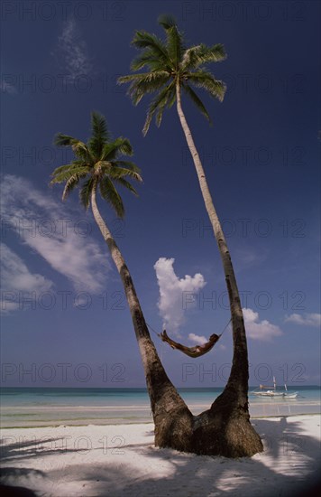 PHILIPPINES, Boracay Island, Man lying in hammock between two coconut palm trees on beach with a sailboat by the shoreline