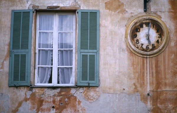 FRANCE, Provence Cotes d’Azur , Alpes-Maritimes, "Sospel.  Architectural detail, clock and window with open green shutters set into outside wall of building with faded, crumbling terracotta plaster."