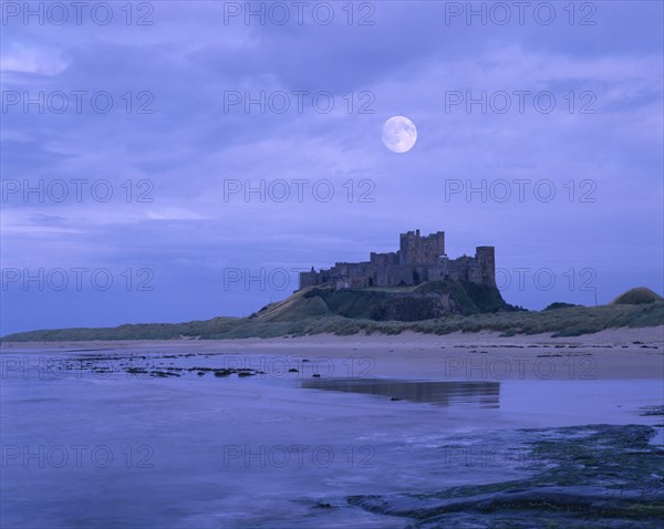 ENGLAND, Northumberland, Bamburgh Castle, View across beach at low tide towards the castle against an early morning sky with the full moon still visible.