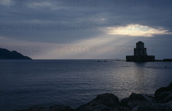 GREECE, Peloponnese, Methoni, View across the sea towards fortress ruins lit by evening sun rays coming through a break in the clouds.