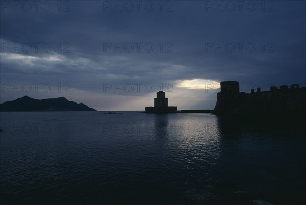 GREECE, Peloponnese, Methoni, View across the sea towards fortress ruins lit by evening sun rays coming through a break in the clouds.