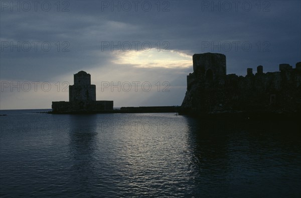 GREECE, Peloponnese, Methoni, View across the sea towards fortress ruins lit by evening sun rays coming through a break in the clouds.
