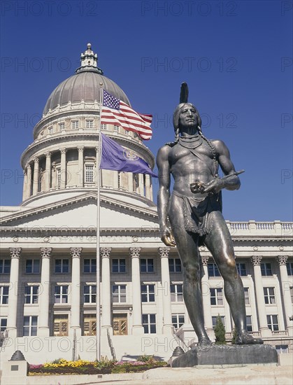 USA, Utah, Salt Lake City, Utah State Capitol.  Detail of dome and colonnaded facade.  Flags flying from flagpole and Indian statue in the foreground.