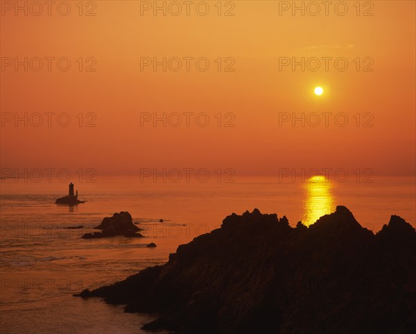 FRANCE, Brittany, Finistere, Near Audierne.  Pointe du Raz at sunset.  Rocks silhouetted against an orange sun and evening sky reflected in the water.