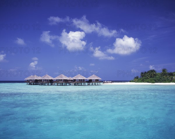 MALDIVES, Baros, "View across expanse of turquoise sea towards thatched beach huts on stilts, beach and tropical vegetation.   "
