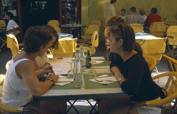 FRANCE, Provence-Cote d’Azur, Bouches-du-Rhône, "Arles.  Le Cafe La Nuit, Place du Forum.  Four young women seated at a table reading a menu. "