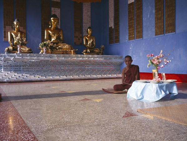 MYANMAR, Yangon, "Shwedagon Pagoda interior. Monk sitting beside a raised altar with three seated, golden Buddha figures. "