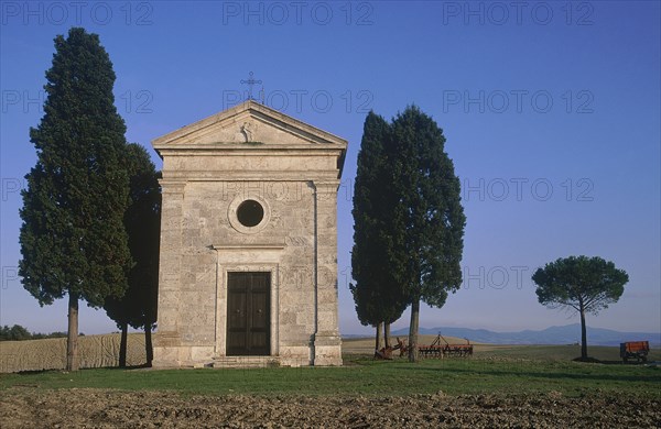 ITALY, Tuscany, San Querico, "Small stone chapel, facade with cypress trees growing beside it in an arable landscape."