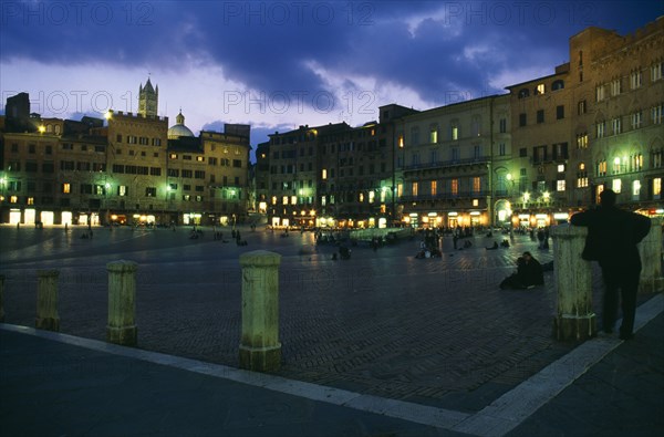 ITALY, Tuscany, Siena, Piazza del Campo. Night view over Il Campo and cafes with people walking or sitting on the red brick paving.