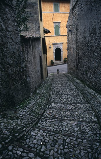 ITALY, Umbria, Spoleto, A narrow cobbled street leading down towards a yellow painted building partly seen between grey stone walls.