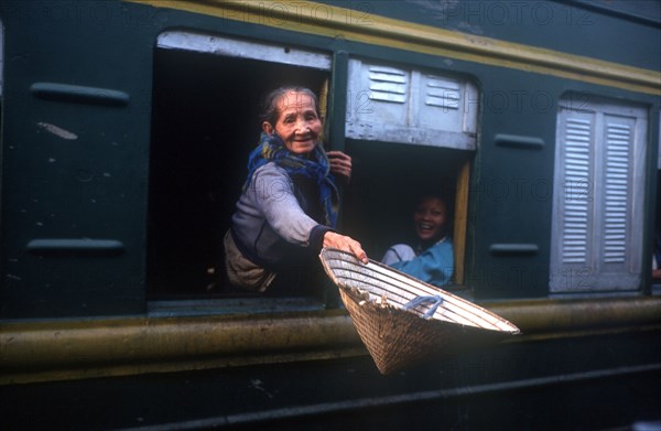 VIETNAM, Hai Van Pass, Old woman reaching out of the window of a train carriage with a straw hat in her outstretched hand.