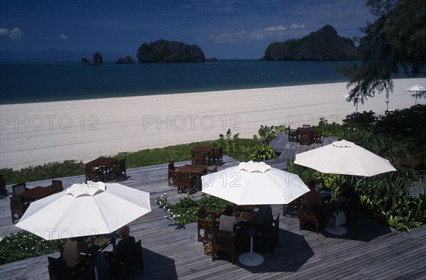 MALAYSIA, Kedah, Langkawi, Tourists sitting under umbrellas set on decking at the garden restaurant of the Radisson Resort hotel on Pantai Rhu beach