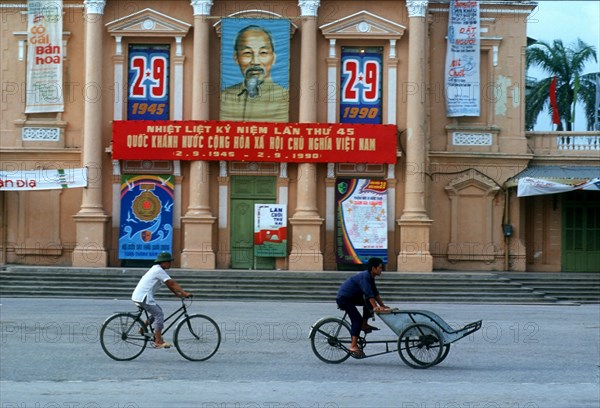 VIETNAM, North, Haiphong, Cyclist and cyclo driver in front of a building draped with commemorative posters of Ho Chi Minh.