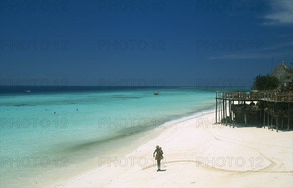 TANZANIA, Zanzibar Island, "Nungwi.  Single figure walking along sandy beach beside aquamarine water.  Beach huts, tables and chairs on raised wooden platform on righthand side."