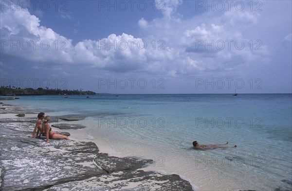 TANZANIA, Zanzibar Island, Mangapwani, Young couple sitting on flat rocks on the shoreline looking out over aquamarine sea with another man lying in the shallow water.