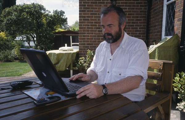 COMPUTERS, Laptop, Man sitting at a table outdoors using a laptop linked to NFT via a mobile phone.