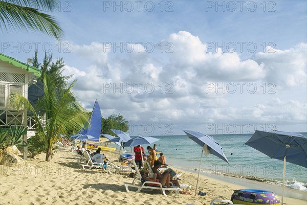 WEST INDIES, Barbados, St Peter, Mullins Bay.  People on loungers with blue sun umbrellas on sandy beach at the waters edge with palm tree and partly seen green and white building on wooden stilts.