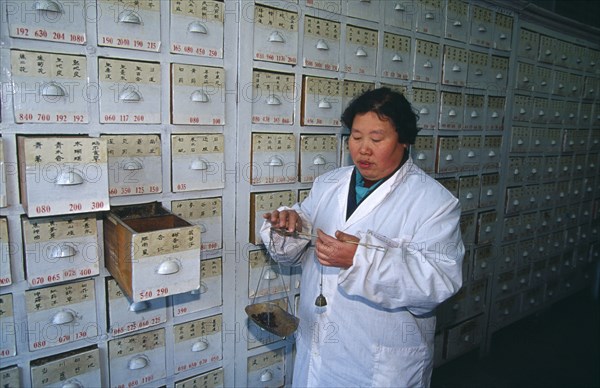 CHINA, Shaanxi Province, Xian, Woman weighing Chinese herbal medicines on hand held scales in front of wall of drawers.