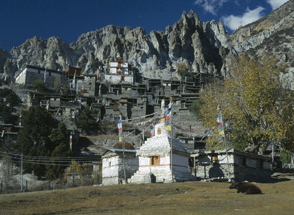 NEPAL, Annapurna Region, Braga, Village near Manang.  Flat roofed stone houses and stupa hung with prayer flags with steep cliffs behind.