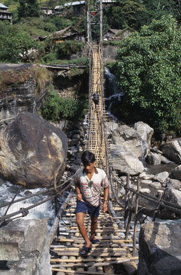 NEPAL, Annapurna Region, Marsyangdi River, Near Besisahar.  Young man crossing wood and rope foot bridge over rocky river gorge.  With person carrying backpack in the opposite direction at far end.