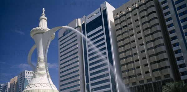 UAE, Abu Dhabi, Al-Ittihad Square. Fountain of water falling from the spout of a sculpted Arabian coffeepot or dallah with high rise buildings behind.