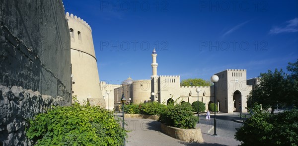 OMAN, Nizwa, "Town centre with crenellated walls surrounding quiet, open, paved area with raised flower beds and trees. "