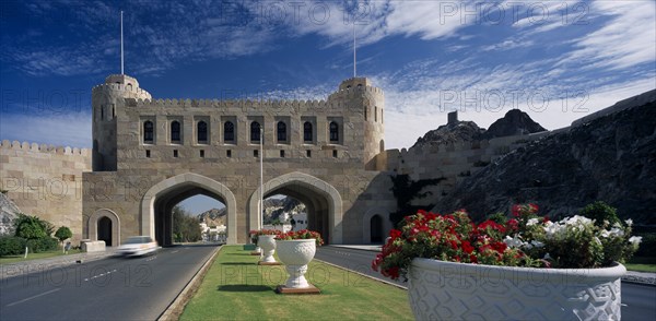 OMAN, Muscat, "Muscat Gate Museum,  Crenellated gatehouse with archways over roads divided by central strip of grass with  white tubs of red and white flowers. "