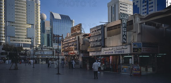 UAE, Abu Dhabi, The Old Souk. Exterior of jewellery and clothes shops with people walking through paved area in front and high rise modern buildings behind.