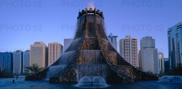 UAE, Abu Dhabi, The Volcano Fountain. Large tiered fountain with water cascading from circular top and high rise modern buildings behind.