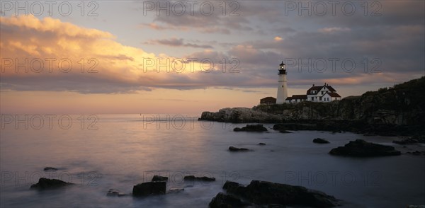 USA, Maine, Portland, Portland Head lighthouse on rocky headland with blue and gold cloudy sky reflected in calm sea.