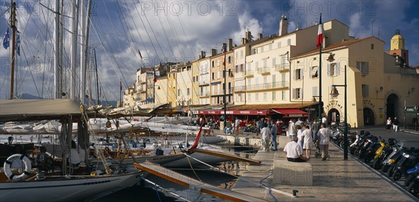 FRANCE, Provence-Cote d’Azur, St Tropez, "View along busy waterfront  with boats moored at the harbourside, open-fronted restaurants and the local tourist office.  Var region."