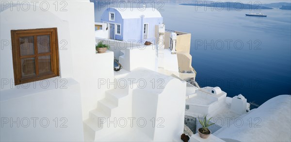 GREECE, Cyclades Islands, Santorini, Oia.  White painted terrace and roof tops overlooking sea with distant ferry and coastline behind.